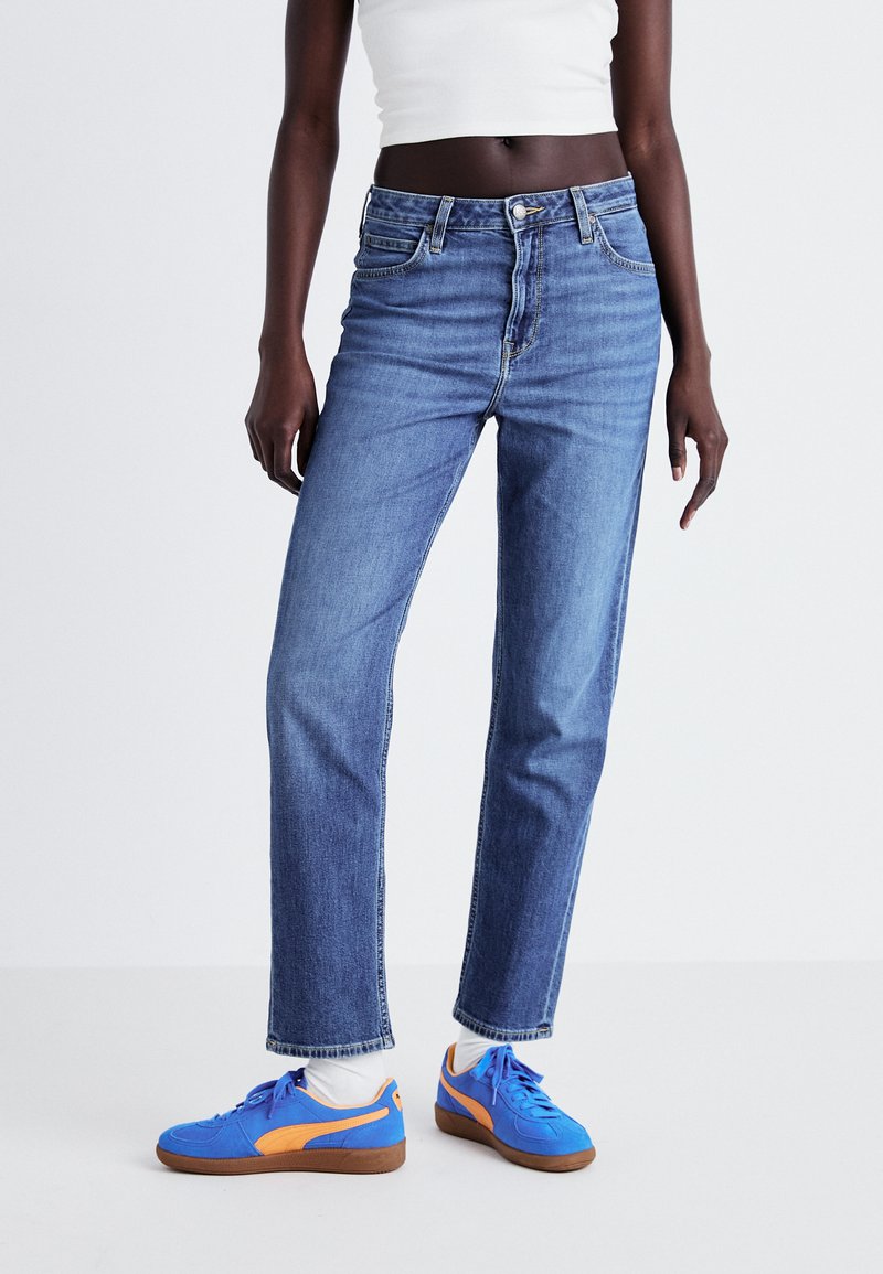 Jean taille haute, jambes droites en denim bleu clair avec cinq poches et une fermeture à bouton, associés à des baskets bleues vives avec des accents orange.