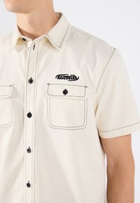 Camicia a maniche corte color crema con cuciture nere, dotata di due tasche frontali e di un logo ricamato sul lato sinistro del petto. Dettagli con bottoni neri.