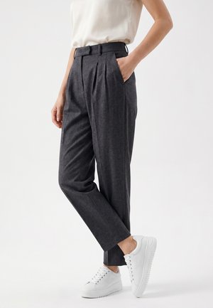 Vrouw draagt hooggetailleerde donkergrijze taps toelopende broek, wit mouwloos topje en witte sneakers, staat met één hand in haar zak.