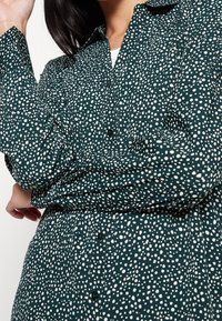 Camicia button-up verde scuro con un motivo a pois bianchi; maniche lunghe con polsini a bottone e un design di colletto classico.