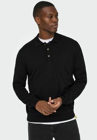 Melns polo džemperis ar garām piedurknēm un apkakli, ar trim pogu aizdares vietām un ribotām aprocēm, valkāts pār baltu T-kreklu.