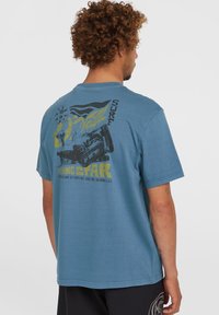 Korte mouwen, blauwe katoenen T-shirt met een grafiek van een surfplank en tekst op de achterkant. Het ontwerp bevat gele en zwarte accenten.