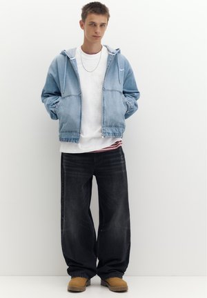 Junger Mann trägt eine blaue Jeansjacke, weißes Hemd, schwarze weite Jeans und braune Stiefel, steht mit den Händen in den Taschen.