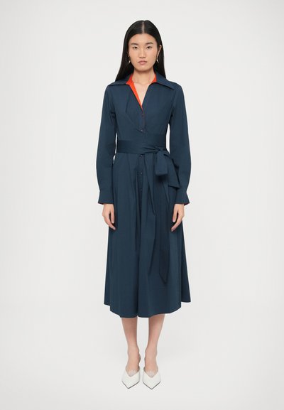 Donkerblauwe jurk met lange mouwen, een tailleband en een V-hals. Heeft een contrasterende rode kraag en knoopdetails, met een vloeiende midilengte rok.