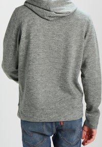 Felpa grigia con cappuccio in un tessuto morbido e testurizzato. Caratterizzata da una vestibilità casual e un orlo a costine, abbinata a jeans blu.