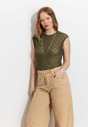 Femme aux cheveux ondulés lâchés portant un haut vert en dentelle sans manches, des colliers argentés superposés et un pantalon beige taille haute à cordon de serrage.