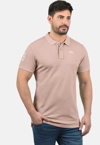 Lichtroze katoenen polo met kraag, een knoopsluiting met twee knopen en een geborduurd logo op de borst. Heeft een nummeraccent op de mouw.
