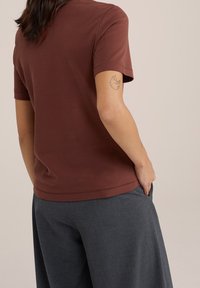 Hnědý krátký rukáv tričko z měkkého materiálu, s kulatým výstřihem a volným střihem. Nositel má minimalistické tetování na horním rameni.