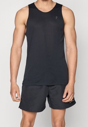 Homme portant un débardeur noir sans manches et un short de sport noir, debout avec les bras détendus contre un fond clair uni.