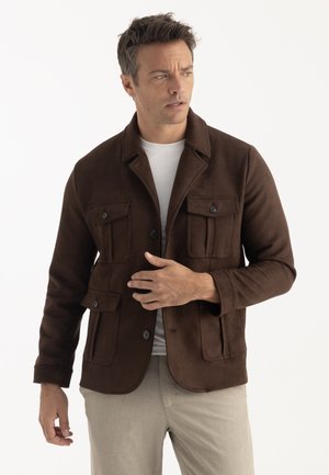 Homme portant une veste marron foncé boutonnée avec des poches à rabat, une chemise blanche en dessous et un pantalon beige clair, regardant vers sa gauche.