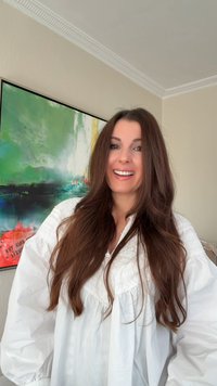 Vit blus med puffärmar och spetsdetaljer, bärs av en person med långt brunt hår, leende framför en färgglad abstrakt målning.