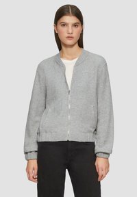 Veste bombardier grise en tissu texturé, avec des poignets et une ceinture côtelés, fermeture éclair à l'avant et deux poches latérales. Portée par-dessus une chemise blanche.