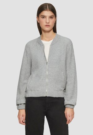 Veste bombardier grise en tissu texturé, avec des poignets et une ceinture côtelés, fermeture éclair à l'avant et deux poches latérales. Portée par-dessus une chemise blanche.