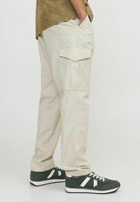 Pantalones cargo beige con bolsillos laterales, corte holgado y material de algodón robusto. Combinados con zapatillas oscuras verdes con acentos blancos.
