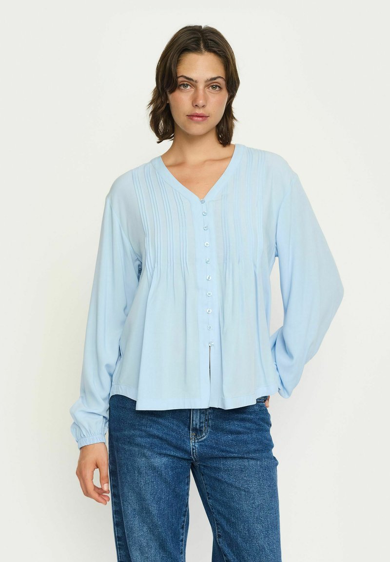 Camicia blu chiaro con bottoni, maniche lunghe, dettaglio frontale plissettato e scollatura a V. Tessuto morbido con una vestibilità comoda. Abbinata a jeans blu scuro.