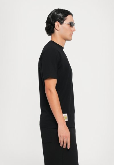 Black short-sleeve T-shirt made of cotton, featuring a round neckline and a small label on the lower side. Model wears slim-fitting black shorts.