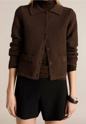 Femme portant un cardigan texturé marron foncé à boutons par-dessus un col roulé marron, associé à un short noir et des bracelets au poignet droit.