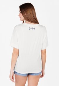 Camiseta de algodón blanca con un corte holgado, mangas cortas y un escote redondeado, que presenta tres pequeños diseños de corazones en la parte superior de la espalda.