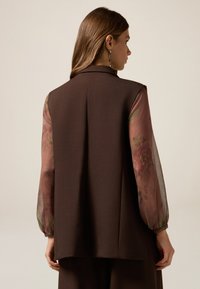 Bruine op maat gemaakte gilet met een hoge kraag, voorzien van sheer bloemenpatroon mouwen in gedempte kleuren. Gladde textuur, eenvoudig ontwerp.