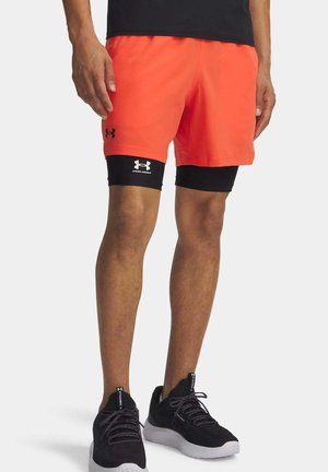 Mann trägt leuchtend orangefarbene Sportshorts mit schwarzen Kompressionslinsen und schwarzen Under Armour Laufschuhen auf weißem Hintergrund.