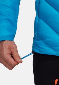 Giacca isolante blu con un motivo a trapunta e un cordoncino elastico all'orlo, mano che regola il cordoncino. Pantaloni corti neri con un accento arancione.