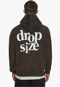 Dunkelbrauner Kapuzenpullover mit großem weißen Text "drop size" auf der Rückseite. Weicher Stoff, Kapuzenentwurf, gerippte Bündchen und Saum.
