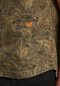 Olijfgroene overhemdbloes met een zwart tropisch bladerenpatroon, voorzien van een oranje geborduurd logo aan de linkerkant. Getextureerde stof.