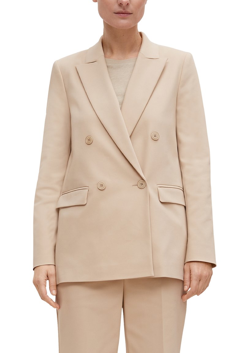 Femme portant un blazer beige croisé avec des poches à rabat et un pantalon assorti, se tenant devant un fond blanc.