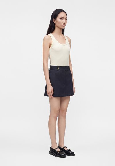 Femme aux longs cheveux foncés portant un haut crème sans manches, une mini-jupe bleu marine foncé avec des fermoirs dorés et des chaussures Mary Jane noires sur un fond uni.