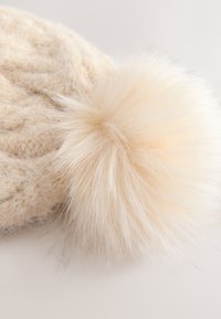 Knitted beige beanie with a large, fluffy cream pom-pom. Soft wool texture, featuring raised patterns on the surface.