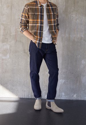 Uomo che indossa una camicia a quadri marrone, arancione e blu sopra una maglietta bianca, jeans blu scuro con risvolti e stivali Chelsea beige, in piedi contro un muro di cemento.