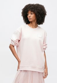 Světle růžová oversize mikina z měkké tkaniny, s kulatým výstřihem, manžetovými rukávy a bílým květinovým motivem na hrudi.