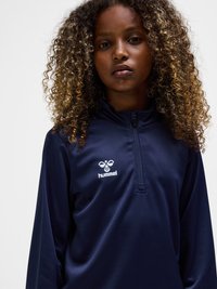 Young person with curly hair wearing a navy blue hummel zip-up sports top, facing forward against a plain background.
