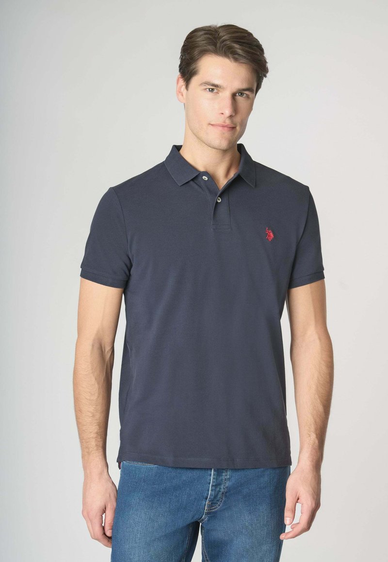 Polo shirt blu navy in cotone, maniche corte, con patta a tre bottoni e un piccolo logo rosso sul lato sinistro del petto. Abbinato a jeans in denim blu.