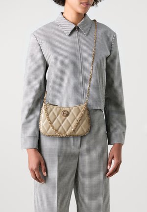Bolso beige acolchado con un logo circular, correa de cadena y textura brillante. Llevado cruzado, combinado con una camisa a cuadros y pantalones de corte tailored.