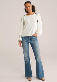 Maglietta bianca con texture a maniche lunghe e ricamo floreale sulle spalle, abbinata a jeans blu chiaro a zampa d'elefante e stivaletti beige.