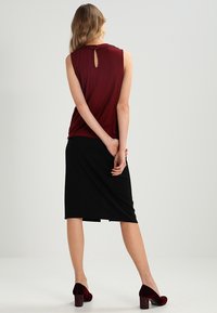 Burgundská bezrukávová blůza s detailem ve tvaru klíče na zádech, v kombinaci s černou sukní do kolen a burgundskými lodičkami s texturovaným povrchem.