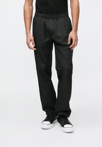 Fekete, könnyű, vízlepergető nadrág elasztikus derékpánttal, egyenes szárakkal és sima textúrával, fekete tornacipővel párosítva.