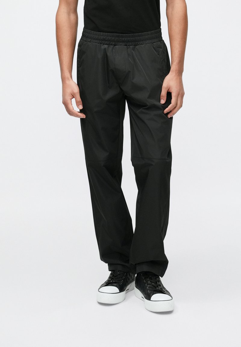 Fekete, könnyű, vízlepergető nadrág elasztikus derékpánttal, egyenes szárakkal és sima textúrával, fekete tornacipővel párosítva.