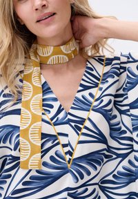 Biała bluzka z niebieskimi wzorami liści, z głębokim dekoltem w kształcie litery V i żółtymi akcentami. Dekolt zdobiony jest żółtym naszyjnikiem z wzorem.