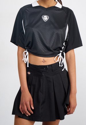 Zwarte cropped sportshirt met witte kraag en zijstrikjes, gecombineerd met een zwarte geplooide rok met hoge taille.