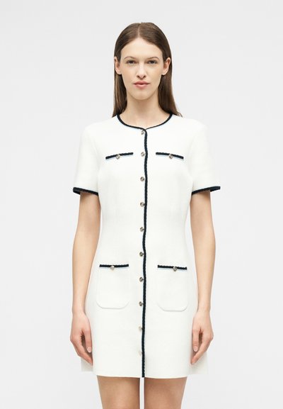 Γυναίκα που φοράει λευκό φόρεμα με κοντά μανίκια και μαύρο περίγραμμα, με κουμπιά μπροστά, δύο τσέπες στο στήθος και δύο χαμηλές τσέπες, πάνω σε απλό φόντο.