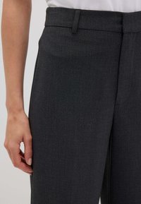 Persona che indossa pantaloni grigi scuri su misura con passanti per cintura, in piedi contro uno sfondo chiaro e uniforme, con la mano rilassata al fianco.