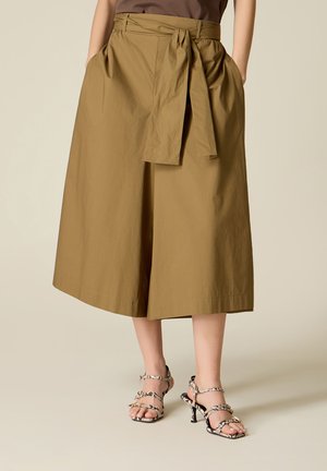 Vrouw draagt een knielange beige rok met een voorstrik en zakken, gecombineerd met sandalen met hoge hakken en een slangenhuidpatroon.
