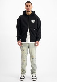 Zwarte zip-up hoodie met een logo aan de voorkant, gecombineerd met lichtblauwe distressed jeans met een relaxed fit en scheuren, gedragen met zwart-witte sneakers.