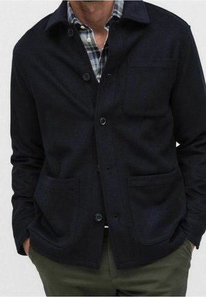 Uomo che indossa una giacca scura blu navy con bottoni sopra una camicia a quadri blu e bianca, pantaloni verde oliva, con le mani nelle tasche.