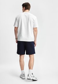 Hvit bomulls T-skjorte med korte ermer, paret med marineblå shorts. Modellen har på seg hvite joggesko og ankelsokker.