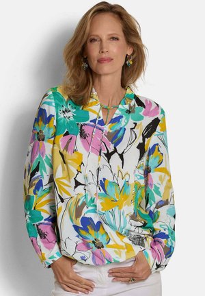 Woman with shoulder-length blonde hair wearing a colorful floral blouse and white pants, with hands resting on hips.