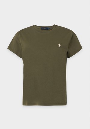 Femme debout portant un t-shirt manches courtes vert olive, un pantalon de jogging noir, des chaussettes blanches et des baskets blanches sur fond blanc.