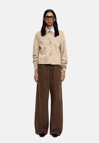 Beżowa zamszowa kurtka z kołnierzem i dwoma przednimi kieszeniami, połączona z szerokimi brązowymi spodniami. Okulary z wyrazistą oprawą dopełniają całość.